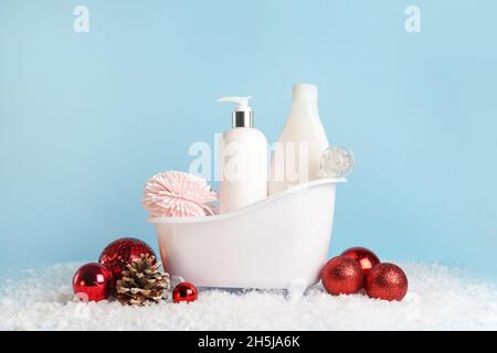 Weiße Badewanne mit Kosmetik, Körperpflegeprodukten und Weihnachtskugeln auf pastellblauem Hintergrund, Winterhautpflege Stockfoto