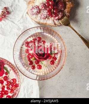 Sektglas mit Rosenblatt und roten Johannisbeeren auf weißem Hintergrund mit Beerentarte. Essen und Trinken Konzept mit roten Früchten, süßem Dessert und champa Stockfoto