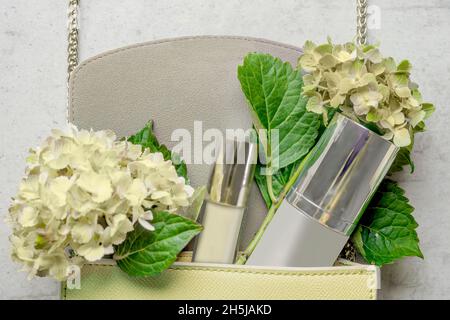 Hellgelbe Handtasche mit Kosmetikflasche, Lipgloss und Hortensien-Blüten auf weißem Holzhintergrund. Feminines Lifestyle-Konzept. Draufsicht. Stockfoto