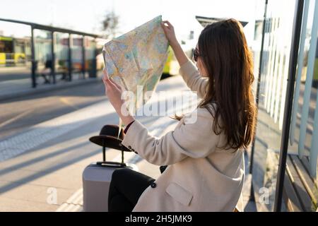 Junge Frau, die an der Bushaltestelle sitzt und den Stadtplan hält. Mädchen schaut sich die Papierkarte an und sucht nach dem Standort, den sie braucht. Reisekonzept Stockfoto