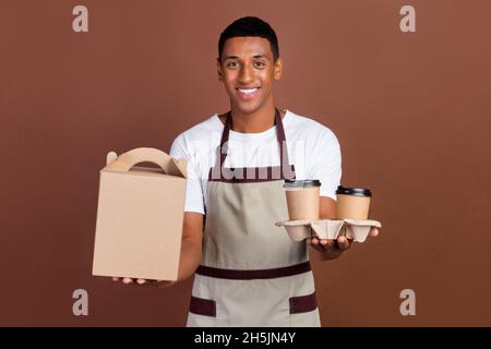Foto von optimistischen Millennial Brunet Kellner Kerl halten Getränke tragen weiße T-Shirt Schürze isoliert auf braunem Hintergrund Stockfoto