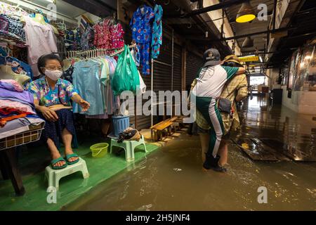 Bangkok, Thailand. November 2021. Eine Frau trägt ihren Sohn, während sie durch die Flut geht, Auf der Seite sitzt ein Ladenbesitzer neben ihrem Laden und hebt ihre Füße über das überflutete Wasser im Wang lang Markt.der Chao Phraya Fluss ist überflutet, beeinflusst durch starke Regenfälle, die Überschwemmungen im Wang lang Markt und in vielen niedrig gelegenen Gebieten entlang des Chao Phraya Flusses verursachen. Kredit: SOPA Images Limited/Alamy Live Nachrichten Stockfoto