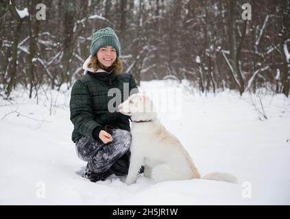 Nettes lächelndes Teenager-Mädchen 16-17 Jahre alt Spaziergänge in verschneiten Wald mit ihrem weißen Hund. Kalte Wintersaison. Aktiver Lebensstil, Wandern mit geliebtem Haustier, jo Stockfoto