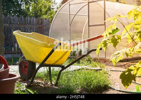 Nahaufnahme eines modernen Polycarbonat-Gewächshauses. Landwirtschaftliche Werkzeuge und ein Wagen mit einem Rad im Garten. Das Konzept des modernen Gartenbaus. Hochwertige Fotos Stockfoto