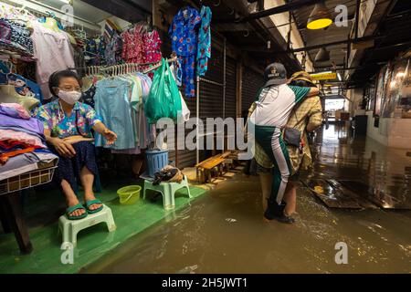 Eine Frau trägt ihren Sohn, während sie durch die Flut geht, Auf der Seite sitzt ein Ladenbesitzer neben ihrem Laden und hebt ihre Füße über das überflutete Wasser im Wang lang Markt.der Chao Phraya Fluss ist überflutet, beeinflusst durch starke Regenfälle, die Überschwemmungen im Wang lang Markt und in vielen niedrig gelegenen Gebieten entlang des Chao Phraya Flusses verursachen. (Foto von Phobthum Yingpaiboonsuk / SOPA Images/Sipa USA) Stockfoto