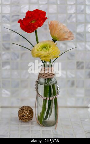 Farbenfrohe Mohn- und Ranunculus-Mohnblumen in einer Glasvase, die mit einer Lederschnur und einer Kette von Strasssteinen umhüllt ist, neben einem Rattanball; Studio Shot Stockfoto