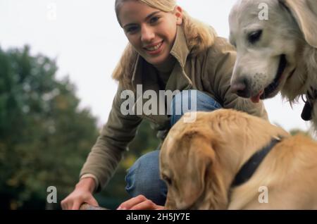 Junge Frau, die sich mit zwei goldenen Retriever-Hunden im Park vor die Kamera beugt; Deutschland Stockfoto