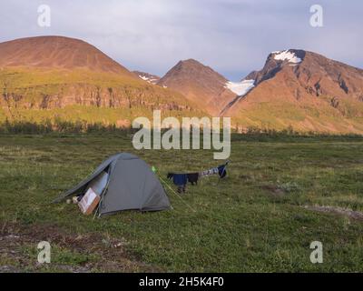 Grünes Trekkingzelt mit hängenden Kleidern in wunderschöner Natur mit Akka-Bergmassiv mit Schnee, Gletscher und Birke in der schwedisch-lappländischen Landschaft Stockfoto