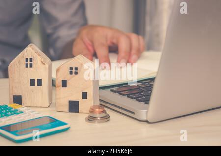 Ein kleines Holzhaus auf dem Schreibtisch EIN Mann Laptop-Computer und Rechner Immobilien-Geschäftskonzept Immobilienversicherung. Stockfoto