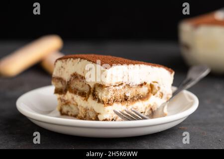 Stück Tiramisu in einem Teller mit Tablett mit Tiramisu und Marienkäfer im Rücken auf dem dunklen Hintergrund. Stockfoto