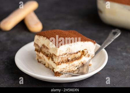 Ein Spitzenfoto eines Stückes Tiramisu mit einer Schale Tiramisu im Rücken mit Marienkäfer auf dunklem Hintergrund. Stockfoto