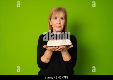 Studioaufnahme einer reifen Dame, die einen Kuchen hält und isoliert auf grünem Hintergrund auf die Kamera schaut Stockfoto