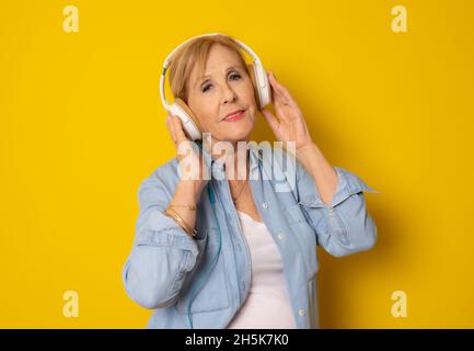 Reife Frau in legerer Kleidung, die Kopfhörer trägt und Musik auf gelbem Hintergrund hört. Stockfoto
