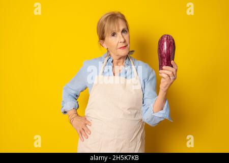 Reife Frau in Schürze, die eine Aubergine isoliert auf gelbem Hintergrund hält. Stockfoto
