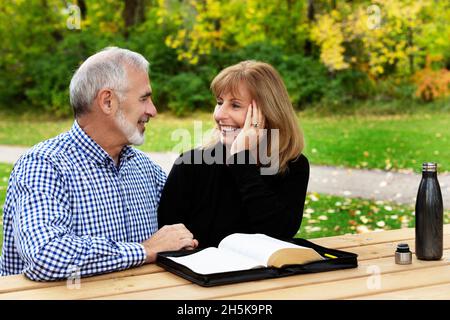 Ein reifes Paar, das sich an einem warmen Herbsttag in einem Stadtpark Zeit mit Hingabe teilt und die bibel an einem Picknicktisch studiert Stockfoto