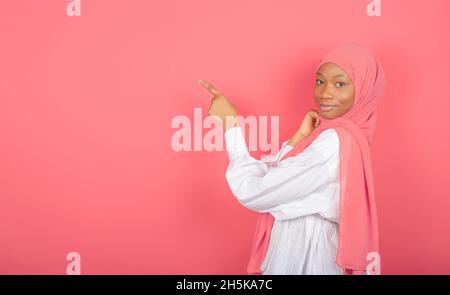 Die junge afroamerikanische muslimische Frau zeigt mit dem Zeigefinger auf den Freiraum, der in einen rosa Schleier gehüllt ist, der auf einem rosa Hintergrund isoliert ist. Stockfoto