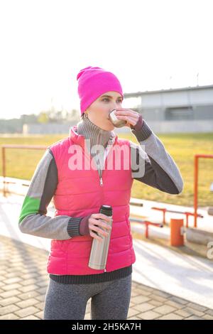 Junge fit Frau in warmen Kleidern hält Metall Thermos trinken heißen Tee oder Kaffee Stockfoto