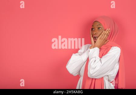 Eine nette junge muslimische Frau berührt ihre Wangen mit beiden Händen, die aufgeregt in den offenen Raum schauen, sie trägt einen rosa Schleier und ein weißes Hemd auf einem rosa Stockfoto