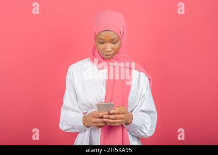 Junge afroamerikanische muslimische Frau mit Smartphone isoliert auf rosa Hintergrund. Junger Mann in rosa Hijab tippt Nachrichten auf Handy. Stockfoto