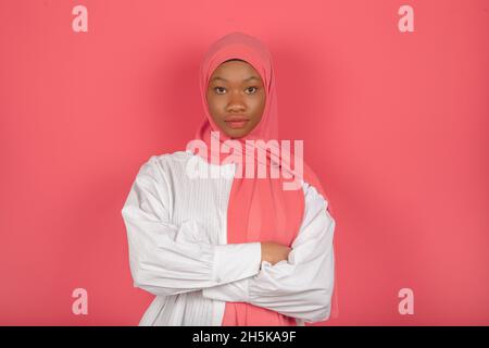 Arabische Frau mit ernsthaftem und ruhigem Ausdruck mit dunkler Haut, eingewickelt in einen rosa Schleier, trägt ein weißes Hemd an der rosafarbenen Studiowand. Stockfoto