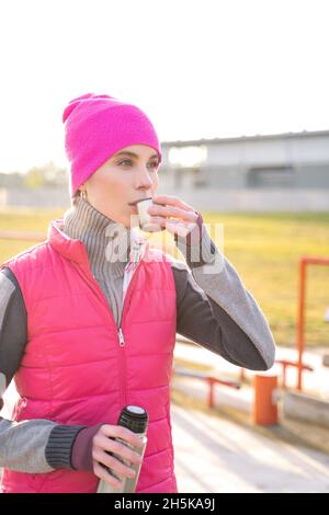 Junge fit Frau in warmen Kleidern hält Metall Thermos trinken heißen Tee oder Kaffee Stockfoto