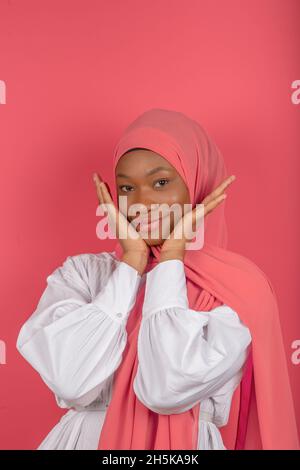 Eine nette junge muslimische Frau berührt ihre Wangen mit beiden Händen mit einem fröhlichen und fröhlichen Blick, trägt einen rosa Schleier und ein weißes Hemd auf einem rosa Hintergrund Stockfoto