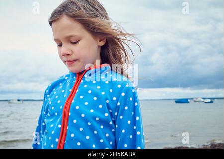 Porträt eines 8-jährigen Mädchens, das an einem bewölkten Tag am Meer steht und nach unten blickt, einige kleine Motorboote im Hintergrund, Ostsee; Deutschland Stockfoto