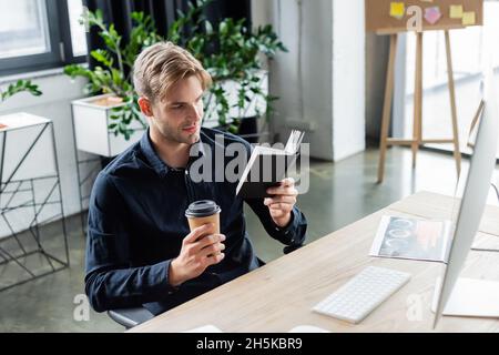 Programmierer hält Papierbecher und Notebook in der Nähe des Computers im Büro Stockfoto