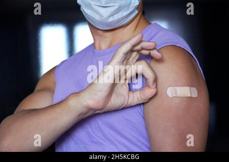 Impfung während der Covid-19-Pandemie, Mann in schützender Gesichtsmaske, der Arm mit Verband zeigt und nach Erhalt des Impfstoffes ein OK-Handzeichen gibt Stockfoto