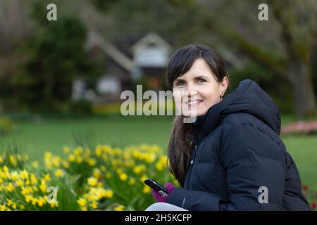 Reife Frau, die in Stanley Park, Vancouver, British Columbia, Kanada, sitzt und ihr Smartphone benutzt Stockfoto
