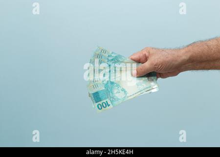 Ältere Männerhand mit rund 100-reais-Banknoten auf blauem Hintergrund. Zahlungskonzept. Hellblauer Hintergrund. Stockfoto