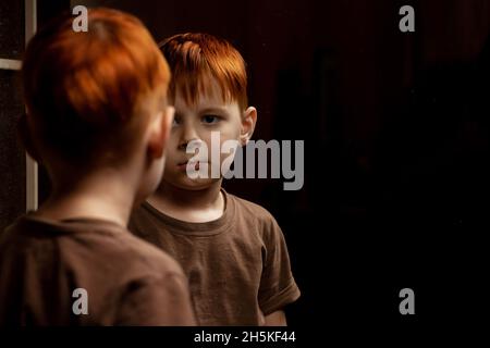 Ein junger rothaariger Junge schaut sich auf dunklem Grund im Spiegel nachdenklich an. Kinderkrise, Depression Konzept. Ort für Ihren Text. Stockfoto