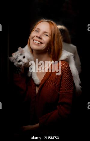 Eine Frau mittleren Alters mit einer weißen Katze auf ihren Schultern lächelt vor einem dunklen Hintergrund. Stockfoto