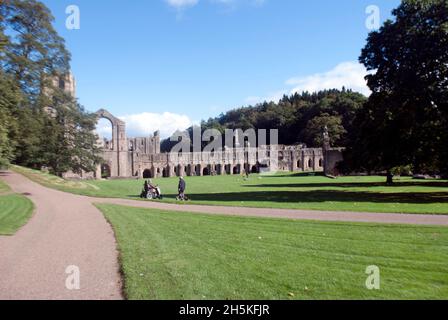 Zwei Personen, die auf dem Gras vor dem Kirchenschiff und dem Großen Kreuzgang der Fountains Abbey, Aldfield, in der Nähe von Ripon, North Yorkshire, England, spazieren Stockfoto