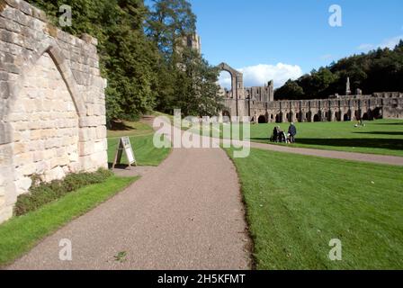 Zwei Personen, die auf dem Gras vor dem Kirchenschiff und dem Großen Kreuzgang der Fountains Abbey, Aldfield, in der Nähe von Ripon, North Yorkshire, England, spazieren Stockfoto