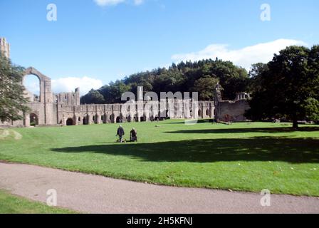 Zwei Personen, die auf dem Gras vor dem Kirchenschiff und dem Großen Kreuzgang der Fountains Abbey, Aldfield, in der Nähe von Ripon, North Yorkshire, England, spazieren Stockfoto