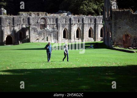 Zwei Personen, die auf dem Gras vor dem Kirchenschiff und dem Großen Kreuzgang der Fountains Abbey, Aldfield, in der Nähe von Ripon, North Yorkshire, England, spazieren Stockfoto