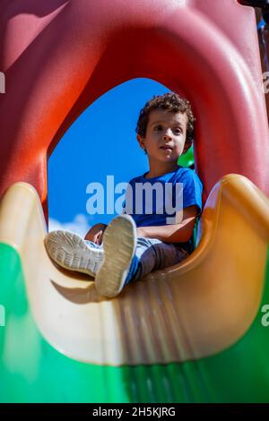 Schöner kaukasischer lockiger Junge, auf dem Spielplatz auf einer Rutsche sitzend Stockfoto