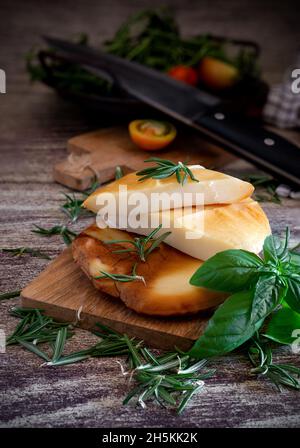 Geräucherte Provola-Medaillons auf Holzschneidebrett mit Kräutern und Gewürzen Stockfoto