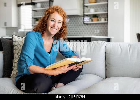 Eine Frau mittleren Erwachsenen sitzt zu Hause auf einer Couch, liest ein Buch und schaut auf die Kamera; Edmonton, Alberta, Kanada Stockfoto