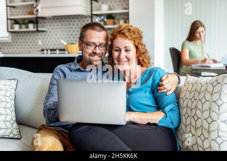 Das Ehepaar sitzt zu Hause auf einer Couch und benutzt seinen Laptop-Computer, während ihre Tochter im Hintergrund Hausaufgaben macht Stockfoto