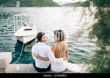 Kotor, Montenegro - 20.06.17: Frisch verheiratetes Paar sitzt umarmt auf dem Pier, an den ein weißes Motorboot vor dem Hintergrund des Meeres gebunden ist Stockfoto