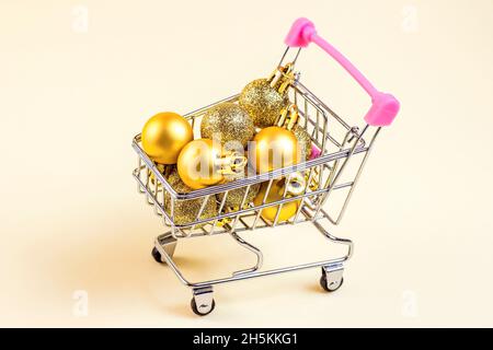 Trolley-Warenkorb mit goldenen Weihnachtskugeln auf hellem Hintergrund. Weihnachtsverkauf, Rabatte, Weihnachtseinkäufe und Geschenkkäufe Konzept. Stockfoto