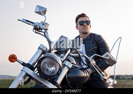 Stilvoller junger attraktiver Mann mit Sonnenbrille sitzt auf einem Motorrad. Porträt eines hübschen jungen Mannes in einer schwarzen Lederjacke am Steuer eines Stockfoto