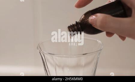 Nahaufnahme eines Medikaments, das aus der Flasche in klares Glas tropft. Konzept. Frau tropft Medizin in Glas auf weißem isoliertem Hintergrund. Behandlung oder Gift Stockfoto