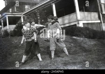 Um 1940, historisch, spielten vier Jungen auf einer Rasenfläche außerhalb der Veranda eines Hauses, wobei die beiden älteren Jungen die beiden jüngeren Jungen auf dem Rücken trugen. Die älteren Jungen tragen Unterhosen oder Hosen, Hosen, die sich knapp unterhalb des Knies erstrecken, und werden mit langen Socken getragen. Auch bekannt als Knickerbocker - oder Golfer plus zwei, da sie sich um zwei Zentimeter unter dem Knie erstrecken - waren diese Baggy-Kneed-Hosen Anfang des 20. Jahrhunderts ein beliebtes Kleidungsstück für Jungen in den USA. Stockfoto