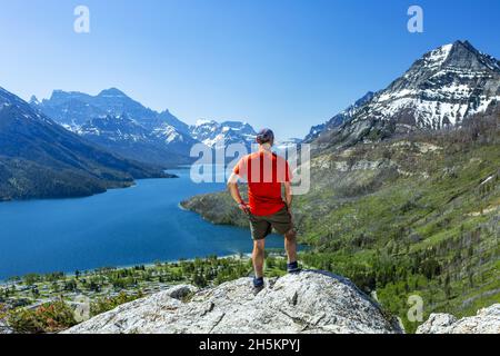 Ein männlicher Wanderer, der auf einer Felsklippe mit Blick auf die Berge und einen See in der Ferne mit blauem Himmel steht, der Waterton Lakes National Park Stockfoto