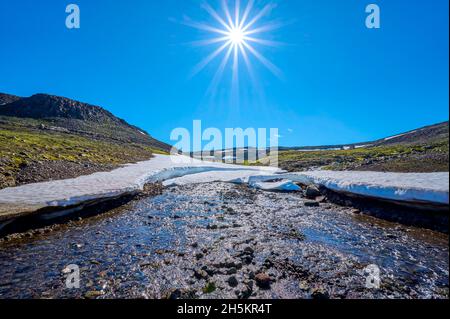 Bergbach mit Schneefeld und Sonneneinbruch in hellblauem Himmel im Sommer, Hellisheidi Eystri Bergpass in Nordisland Stockfoto