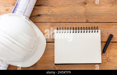 Büroschreibtisch des Architekten-Ingenieurs, leeres Notizbuch, Bauzeichnungen und weißer Schutzhelm auf einem Holztisch. Draufsicht, Platz Stockfoto