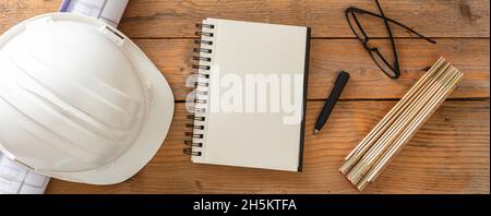 Büroschreibtisch des Architekten-Ingenieurs, leeres Notizbuch, Bauzeichnungen und weißer Schutzhelm auf einem Holztisch. Draufsicht, Banner Stockfoto